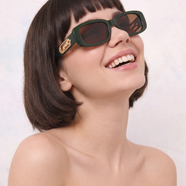 Woman wearing dark green oversized sunglasses, smiling with bangs and an off-the-shoulder white top on a light background.