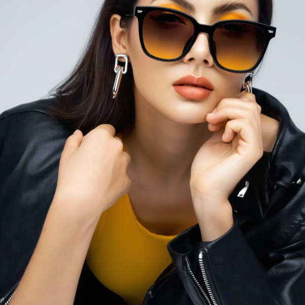Woman wearing oversized black sunglasses, a yellow top, and a black leather jacket, posing with hands near her face.