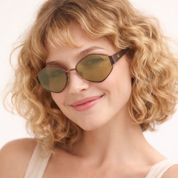 Woman with curly blond hair wearing green-tinted sunglasses and a beige top, smiling softly at the camera.