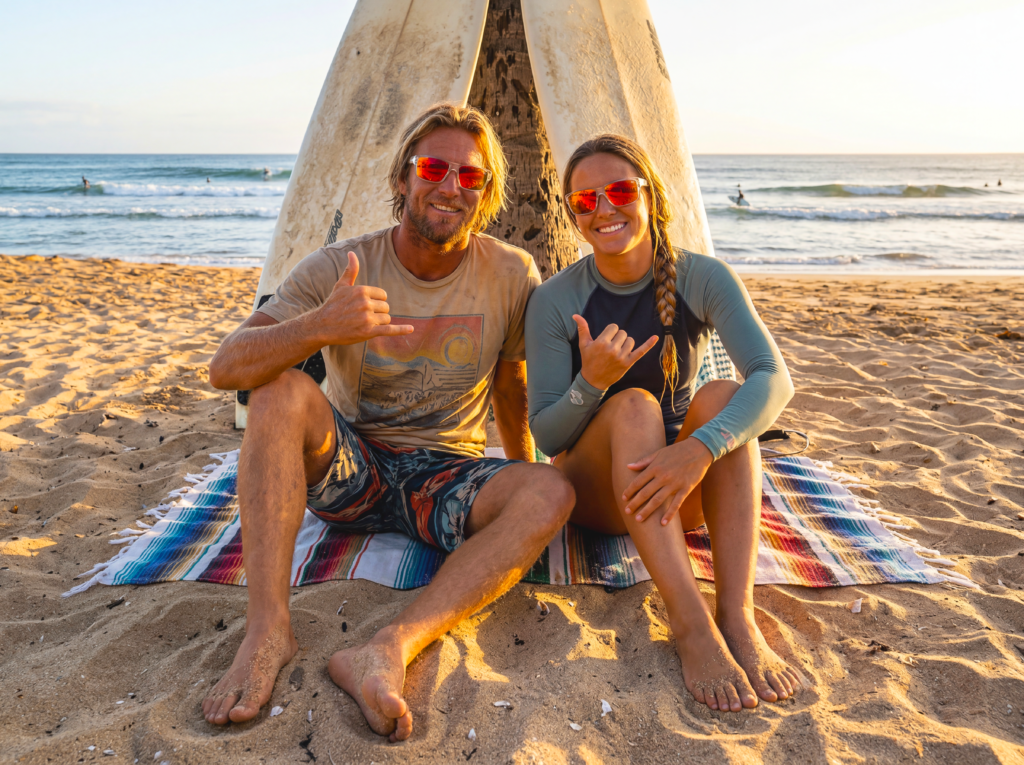 Man and woman wearing sunglasses sit on a striped towel on the beach, giving thumbs up with a surfboard propped between them.
