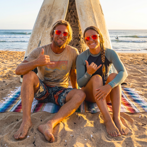 Man and woman wearing sunglasses sit on a striped towel on the beach, giving thumbs up with a surfboard propped between them.