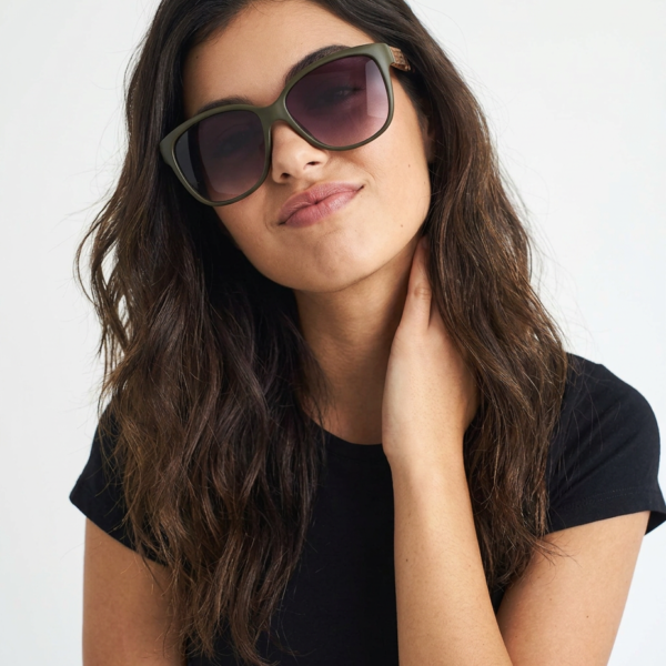 Woman wearing oversized olive-green sunglasses and a black t-shirt, tilting her head slightly and touching her neck with one hand.