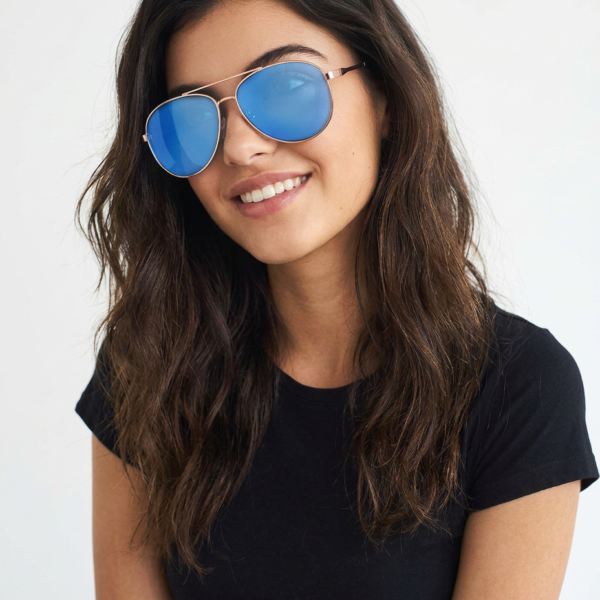Woman with wavy brown hair wearing blue mirrored sunglasses and a black t-shirt, smiling at the camera.