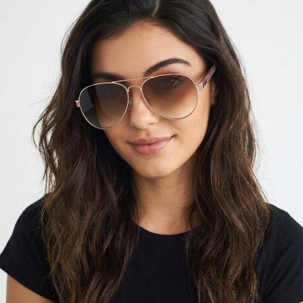 Woman with wavy brown hair wearing aviator-style sunglasses and a black shirt, looking at the camera against a light background.