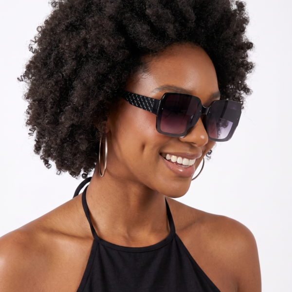 Woman with a voluminous curly afro, wearing oversized sunglasses and large hoop earrings, smiling against a light background.