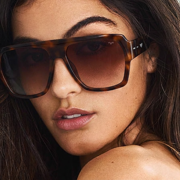 Close-up of a woman with long dark hair wearing oversized tortoiseshell sunglasses, lips slightly parted.