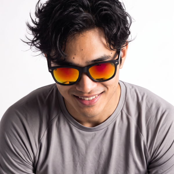 Smiling man with dark wavy hair wearing black sunglasses with orange mirrored lenses, gray shirt, close-up portrait from chest up.