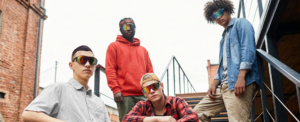 Four young men pose on an outdoor metal staircase, wearing casual streetwear and reflective sunglasses.