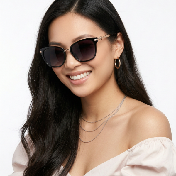 Smiling woman with long dark hair wearing oversized black sunglasses, an off-the-shoulder pale pink top, hoop earrings, and layered silver necklaces.