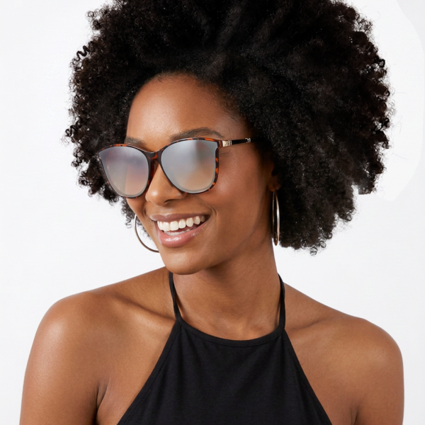 Smiling woman with a large curly afro, wearing tortoiseshell oversized sunglasses, hoop earrings, and a black halter top against a white background.